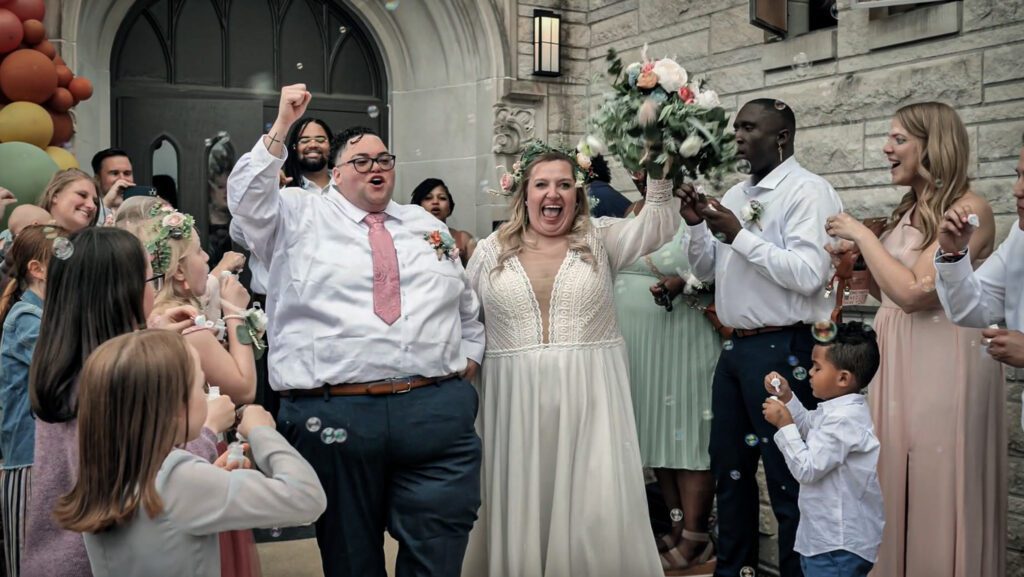 Image of a masculine AFAB Non-binary person and their femme wife walk through a bubble exit, surrounded by their cheering friends, with arms raised in celebration of their new marriage. The couple is white and plus size, and their friends are a wide mixture of races and sizes.