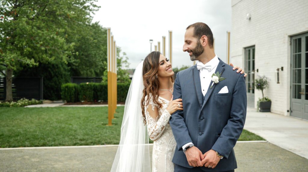 A hetero Arab couple looks at each other, the bride stands behind the groom and looks up at him with a smile. She wears a lace dress with a long veil, and she has long curly hair. The groom stands taller in a dusty blue suit, smiling at his wife