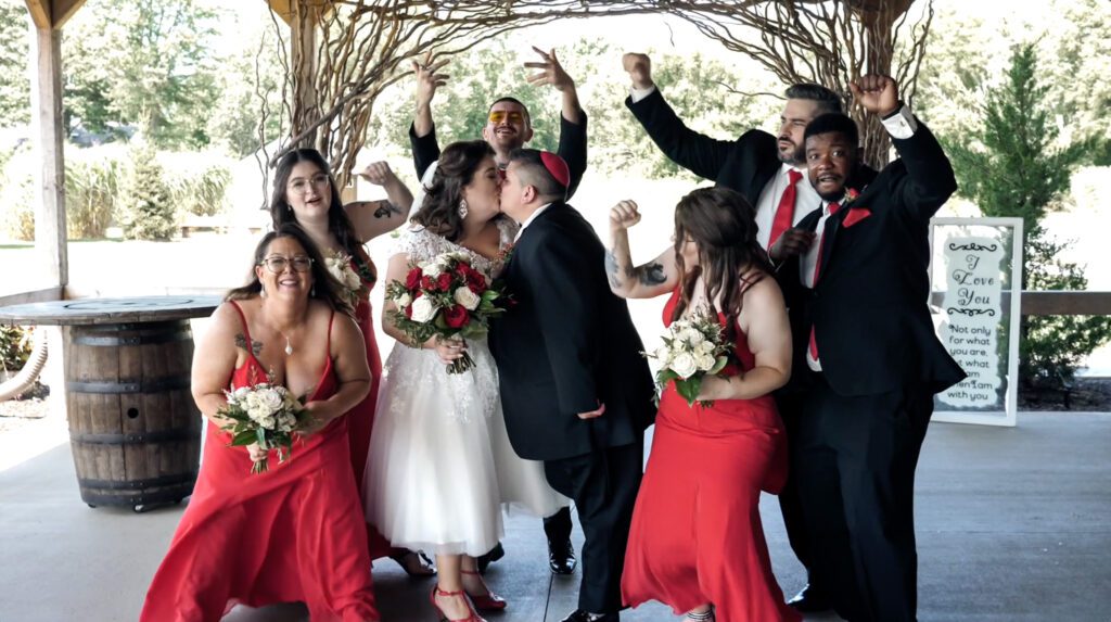 Image of a jewish lesbian couple kisses in the center of the frame, surrounded by their cheering friends. The groups is on the deck of a winery wearing wedding attire with red accents. 