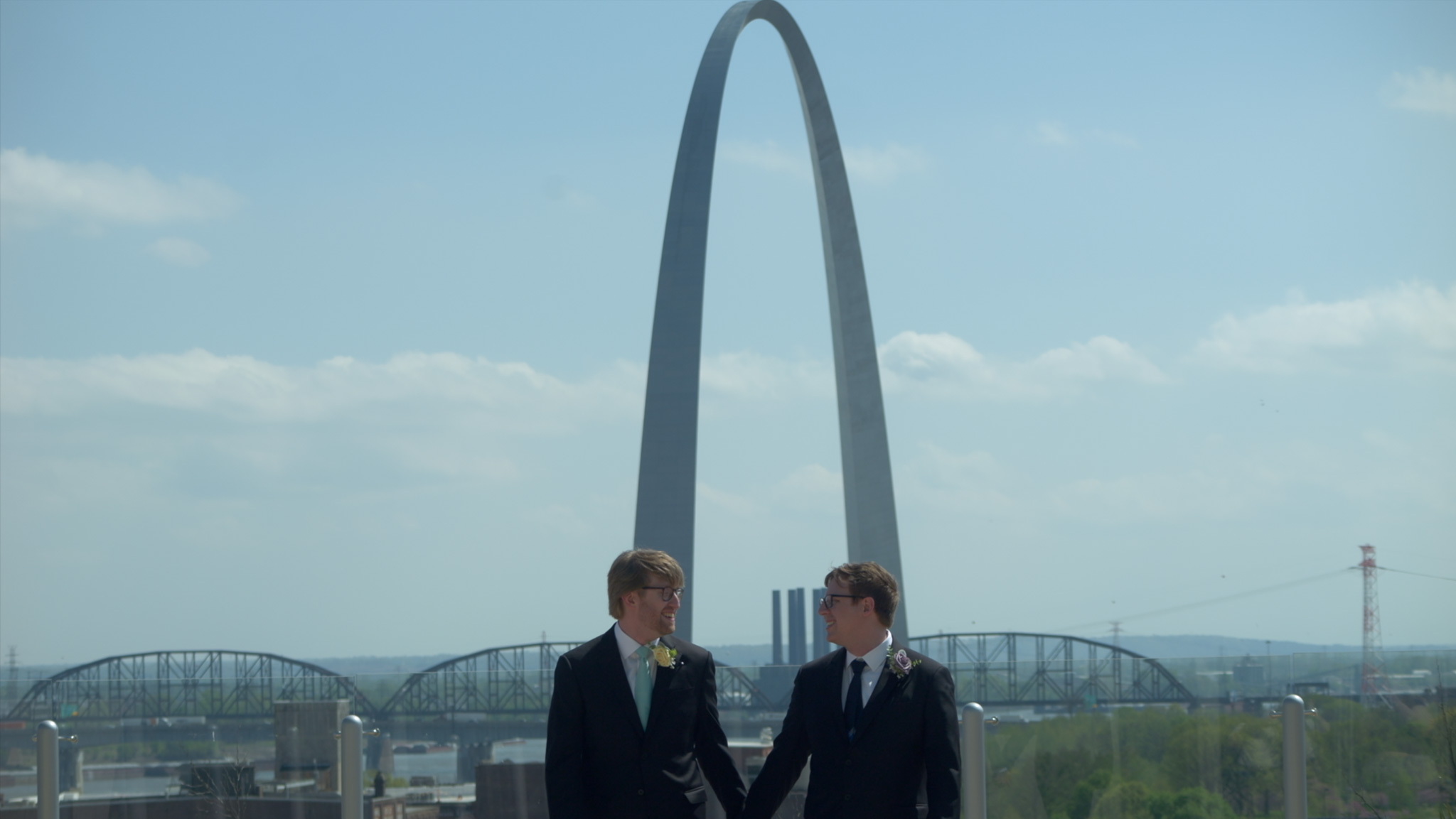Two white gay men hold hands at their wedding, walking toward the camera with a large blue sky and the St. Louis arch in the background.