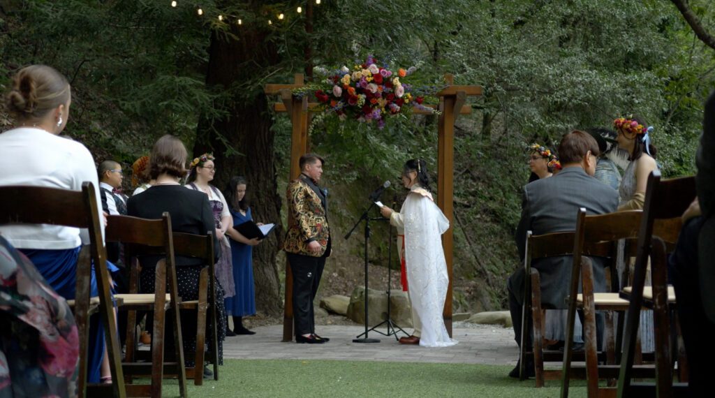Kat and Bear stand at their altar, a wooden arch with bright, warm florals outside in the redwood forest. Bear is a white non-binary person with short brown hair, a gold and red paisley suit jacket and black slacks. Kat, is an asian non-binary person wearing an eastern-inspired white cape with red accents, and a cross-body white robe tied at the waist with a red cord. 